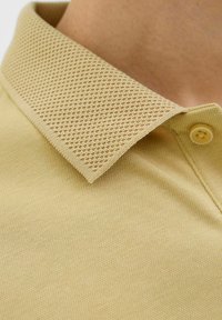 Šviesiai smėlinės spalvos polo marškiniai su tekstūruotu tinkliniu apykakle, vienoduoju korpusu ir vienu mygtuku apykaklėje.