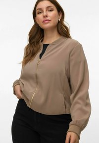 Veste bomber courte beige avec une texture lisse, fermeture éclair dorée, poignets côtelés et poches latérales. Portée par-dessus un haut noir et associée à un pantalon foncé.