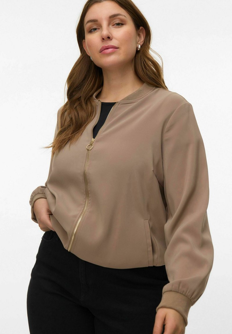 Veste bomber courte beige avec une texture lisse, fermeture éclair dorée, poignets côtelés et poches latérales. Portée par-dessus un haut noir et associée à un pantalon foncé.