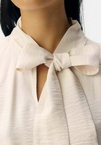 Cremfarbene Bluse aus strukturiertem Stoff mit einem Schleifen-Detail am Kragen und einem dezenten Schlitz, der das elegante Design unterstreicht.