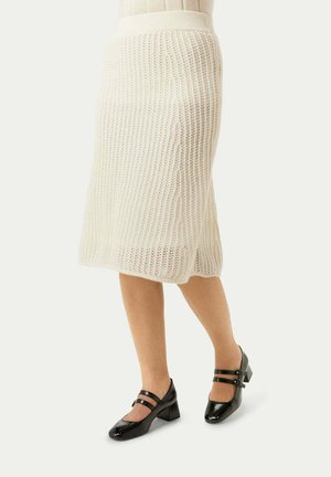 Jupe midi blanche en tricot avec taille côtelée, présentant un motif texturé. Associée à des chaussures Mary Jane en cuir verni noir.