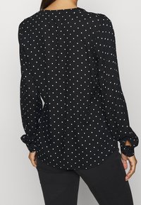 Personne portant un chemisier noir à manches longues avec des pois blancs, vu de dos, associé à un pantalon noir.