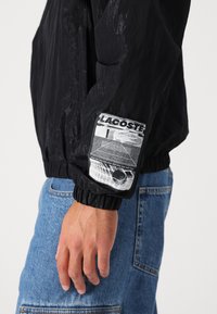 Jachetă neagră din material waterproof, cu o textură specială, manșete elastice și un patch grafic cu "LACOSTE" pe mâneca stângă.