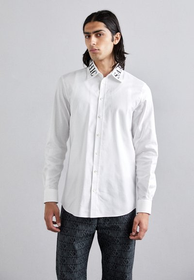 Camicia bianca con bottoni, con testo ricamato in nero sul colletto, abbinata a pantaloni neri con un motivo a logo ripetitivo.