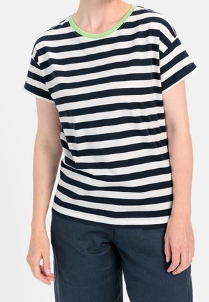 Persona que lleva una camiseta de manga corta con rayas horizontales en blanco y negro, con escote verde y pantalones oscuros, de pie con los brazos relajados.