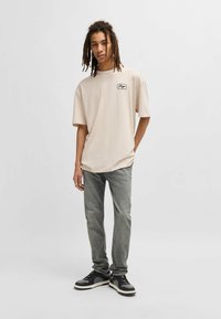 Beige oversized t-shirt med en liten logotypplåt, matchad med blekta grå jeans och svarta sneakers med vita detaljer.