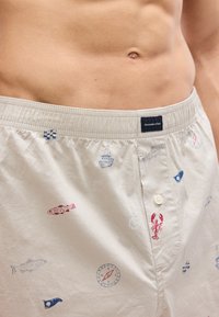 Ανδρικός κορμός που φοράει μπεζ ανδρικά μποξεράκια με ναυτικά σχέδια, συμπεριλαμβανομένων ψαριού, αστακού, βάρκας, πυξίδας και σημαιών, ελαστική μέση με ετικέτα μάρκας.