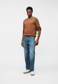 Uomo che indossa un maglione marrone a maniche lunghe, jeans blu e sneakers bianche, in piedi davanti a uno sfondo grigio chiaro uniforme.