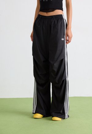 Pantalons de sport noirs avec des bandes blanches sur les côtés, coupe ample, taille élastique et cordon de serrage. Associés à des chaussures jaunes.