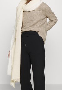 Pull beige en tricot à coupe ample, associé à un pantalon noir fuselé. Une écharpe douce de couleur claire est drapée autour du cou. Détails de tissu texturé.