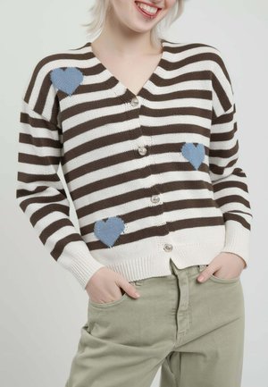 Persona che indossa un maglione a righe marroni e bianche con bottoni e motivi a cuore blu, mani nelle tasche di pantaloni verde chiaro, sorridente.