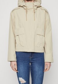 Beige hættejakke med snoretræk og lommeklapper med knapper, båret over blå slidte skinny jeans på en neutral baggrund.