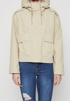 Beżowa kurtka z kapturem, z troczkami i zapinanymi na guziki kieszeniami, носzona на неbieských, с tornadi skinny джырха на нейтральном фоне.