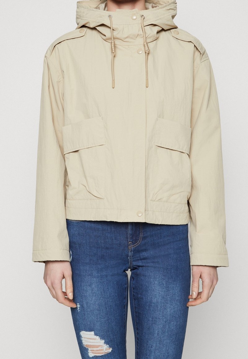 Beige hættejakke med snoretræk og lommeklapper med knapper, båret over blå slidte skinny jeans på en neutral baggrund.