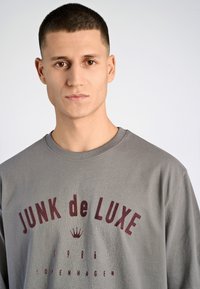 Sudadera de algodón gris con estampado "JUNK de LUXE" en burdeos, cuello redondo y corte relajado. Diseño simple con banda en el cuello de canalé.