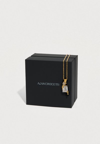 Κολιέ με χρυσό μενταγιόν με διαυγή ορθογώνιο πέτρα και μικρότερα διακοσμητικά, εκτεθειμένο σε μαύρο κουτί με λογότυπο.