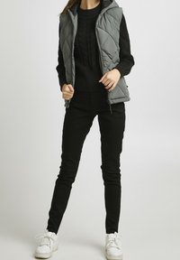 Grå vadderad väst med huva över en svart kabelstickad tröja, kombinerad med svarta slim fit-byxor och vita sneakers.