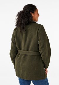 Cappotto di lana verde oliva con vita segnato da una cintura e una vestibilità leggermente oversize. Caratterizzato da un colletto classico e un tessuto texturizzato.