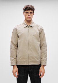 Beżowa kurtka zapinana na zamek z ukośnym kołnierzem i haftowanym logo. Posiada długie rękawy oraz gładką, matową teksturę. Czarne dżinsy dopełniają stylizację.