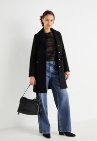 Abrigo de lana negro con botones dorados, suéter de punto multicolor, jeans de denim de pierna ancha y un bolso negro. Zapatos negros de tacón añaden contraste.