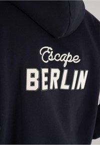Navy blue hoodie with embroidered white text "Escape" in script and large block letters "BERLIN" on the back under the hood.