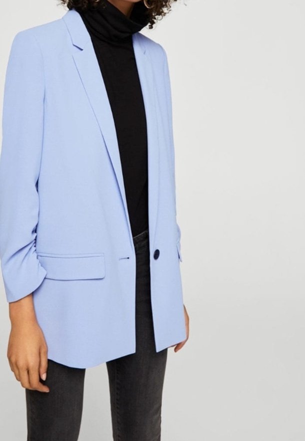 Personne portant un blazer bleu clair sur un col roulé noir et un jean gris foncé, debout devant un fond clair uni.