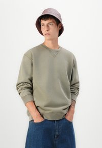 Sudadera verde oliva con cuello redondo y puños acanalados, combinada con pantalones de mezclilla azul. La persona lleva un sombrero de cubo rosa claro.