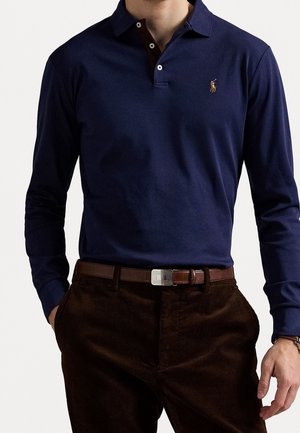 Mann trägt ein navyblaues Poloshirt mit langen Ärmeln und kleinem Logo sowie dunkelbraune Hose mit Gürtel, Hände in den Taschen, vor weißem Hintergrund.