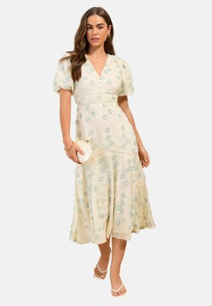 Frau in einem cremefarbenen Midi-Kleid mit blauer Blumenstickerei, Puffärmeln, die eine runde weiße Clutch hält, weiße Sandalen und goldenen Schmuck trägt.