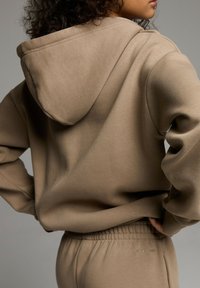 Beżowa bluza z kapturem w luźnym fasonie, wyposażona w ściągacz przy kapturze, prążkowane mankiety i gładką teksturę. Połączona z dopasowanymi spodniami dla uzyskania spójnego wyglądu.