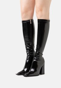 Bottes noires montantes jusqu'au genou avec une finition vernissée brillante, coupe slim, bout pointu, et talon large. Équipées d'une fermeture éclair latérale pour un enfilage facile.
