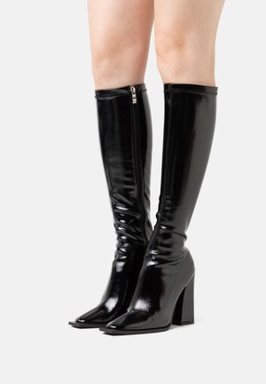 Bottes noires montantes jusqu'au genou avec une finition vernissée brillante, coupe slim, bout pointu, et talon large. Équipées d'une fermeture éclair latérale pour un enfilage facile.