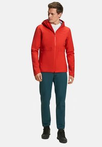 Rote wasserdichte Jacke mit Kapuze, Reißverschluss und Seitentaschen. Kombiniert mit türkisfarbenen Hosen und schwarzen Schuhen für den Außenbereich.