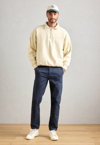 Mann trägt einen beigen Sweatshirt mit Viertelreißverschluss, marineblaue Hose, weiße Sneakers und eine weiß-grüne Kappe. Er steht mit den Händen in den Taschen auf einem Holzboden.