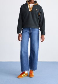 Pullover en polaire gris foncé avec col et boutons beige, associé à un jean bleu à jambes larges et des baskets orange avec des rayures vertes.