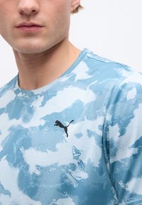 Κοντινό πλάνο ενός ατόμου που φοράει μπλε ανοιχτό και λευκό πουκάμισο με σχέδιο της Puma και μαύρο λογότυπο Puma στο στήθος.