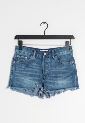 Shorts en denim bleu clair avec un ourlet effiloché, design à cinq poches et rivets en cuivre. Ils sont accrochés à un cintre en métal noir sur fond blanc.