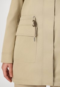 Cappotto beige con una texture liscia, dotato di una tasca laterale, dettagli con cordoncino e cuciture a contrasto lungo l'orlo e le maniche.