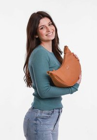 Hnědá kožená měsíční taška s texturovaným povrchem, se znakem a zaobleným designem. Model má na sobě žebrovaný svetr v barvě smaragdové a ošoupané džíny.