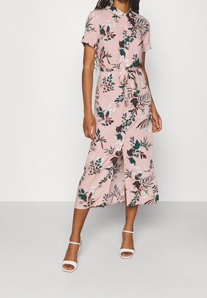 Frau trägt ein pinkes Midikleid mit grünen und weißen Blumenmustern, kurzen Ärmeln, Knopfleiste vorne, Taillengürtel und weißen offenen High Heels.