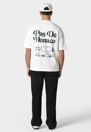 Hvid t-shirt med korte ærmer og sort grafisk tekst "Pas De Monaco" samt bilillustrationer på ryggen; kombineret med sorte bukser.