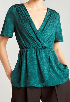 Tyrkysová halenka ve stylu zavinovacího střihu s jemným květinovým vzorem, krátké rukávy, nařasený pas, v kombinaci s tmavě hnědými kalhotami, ruce v kapsách.