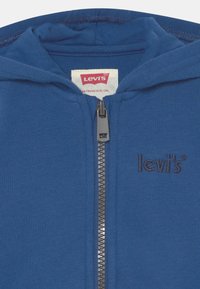 Modrá mikina na zip s vyšitým logem Levi's na hrudi a značkovou etikettou Levi's uvnitř límce.
