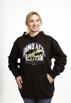 Uśmiechnięta kobieta o blond włosach, ubrana w czarną bluzę z kapturem z wizerunkiem ryczącego dinozaura i napisem "Dino Ace Racing Team Atlanta Speedway."