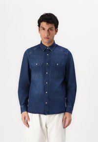 Camisa de mezclilla azul oscuro con mangas largas, cierre delantero de botones, dos bolsillos en el pecho y un cuello estándar. Tejido de algodón con una textura suave.
