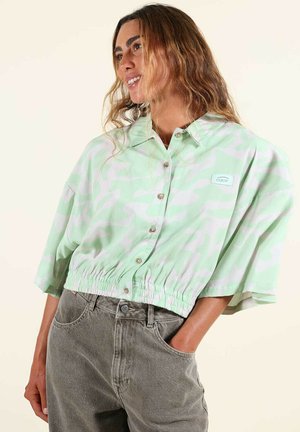 Femme aux longs cheveux ondulés portant un haut court boutonné à manches courtes vert clair et blanc avec une taille élastique, et un jean taille haute gris.