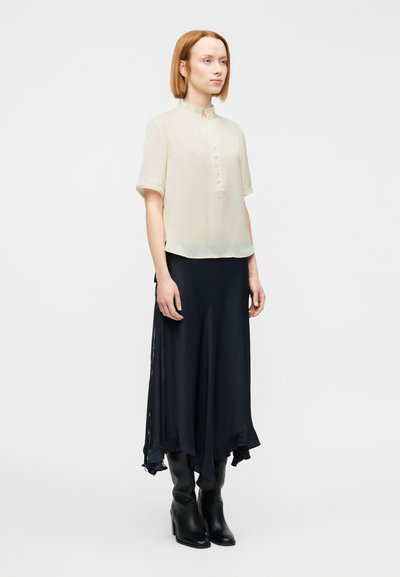Ανοιχτό μπεζ μπλούζα με κοντό μανίκι και κινέζικο γιακά, με λεπτομέρεια κουμπιών, σε συνδυασμό με μια ροή μαύρη φούστα και μαύρες μπότες μέχρι το γόνατο.