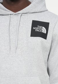 Felpa grigia realizzata in tessuto morbido, caratterizzata da un grande logo quadrato nero con testo bianco e un accento curvo, insieme a una tasca a marsupio.