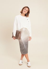 Weißer Pullover mit strukturiertem Design und Knöpfen, kombiniert mit einem silbernen, mit Pailletten besetzten Midirock. Die Accessoires umfassen eine braune Tasche und weiße Sneaker.