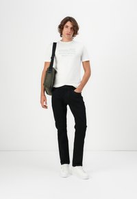 Camiseta blanca con texto impreso, jeans negros y zapatillas blancas. El modelo sostiene un bolso verde oliva con correa de hombro.